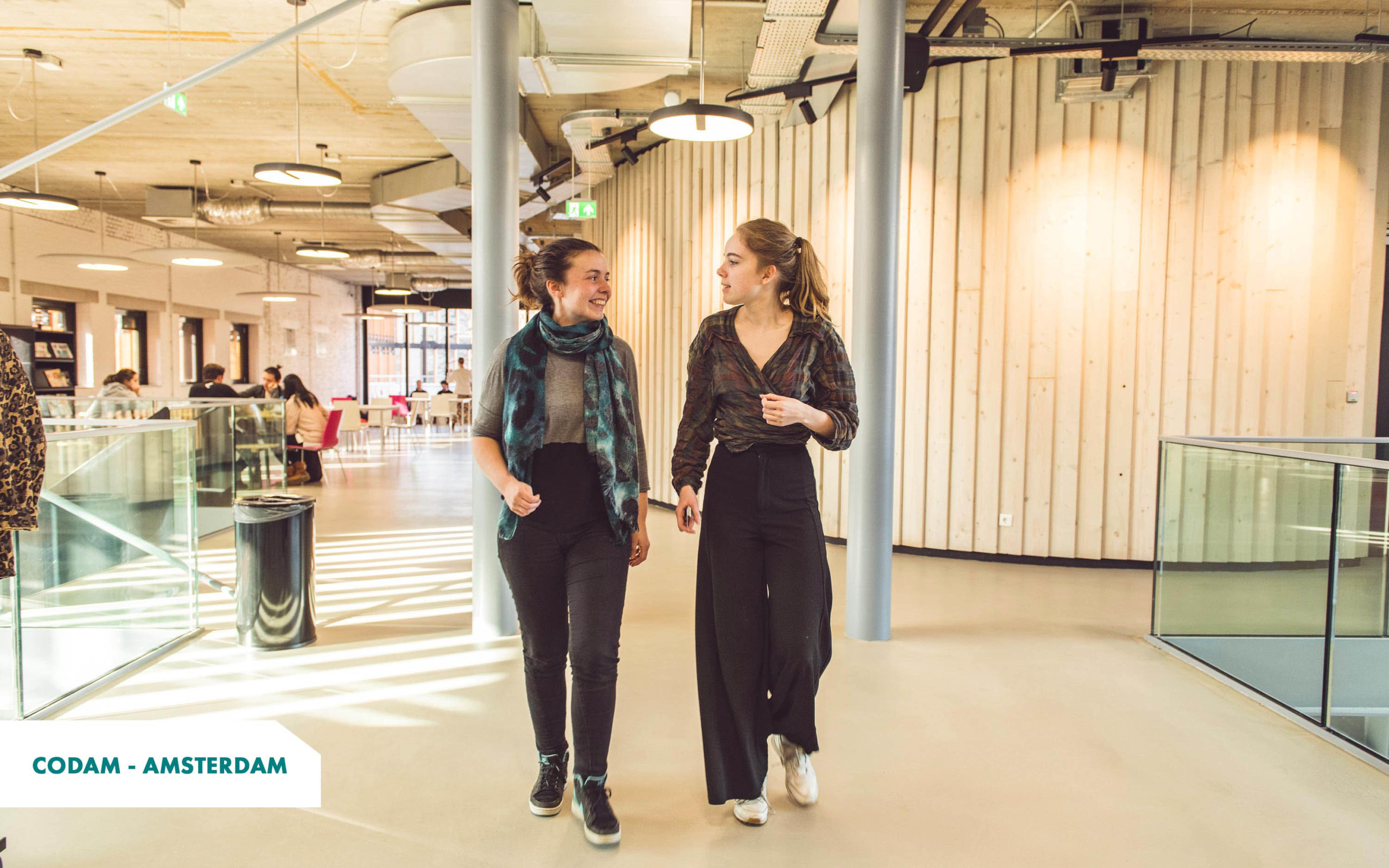 2 women talking while walking in the lighted corridors of Codam's campus (The Netherlands)